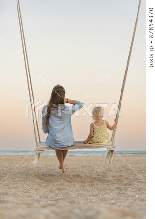 Mom and little girl sit on swing and watch the stunning sunset over the sea. Magical sunrise over sea. Mom and little girl sit on swing and watch the stunning sunset over the sea. Magical sunrise over sea. 87854370
