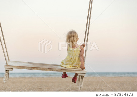 Cute little blonde girl gets on the swings on the beach on sea background. Holidays with children on the sea. Magical light of sunset Cute little blonde girl gets on the swings on the beach on sea background. Holidays with children on the sea. Magical light of sunset 87854372