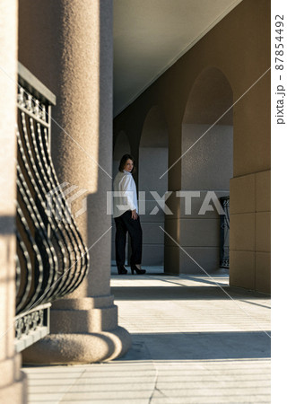 Woman stands hal turn near column of beautiful building. Full-length portrait of girl in trouser suit. Woman stands hal turn near column of beautiful building. Full-length portrait of girl in trouser suit. 87854492