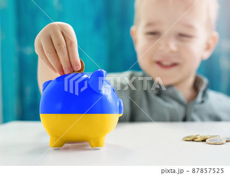 Child sitting at the table with money coins and biggybank in colour of Ukranian flag. Child sitting at the table with money coins and biggybank in colour of Ukranian flag. 87857525