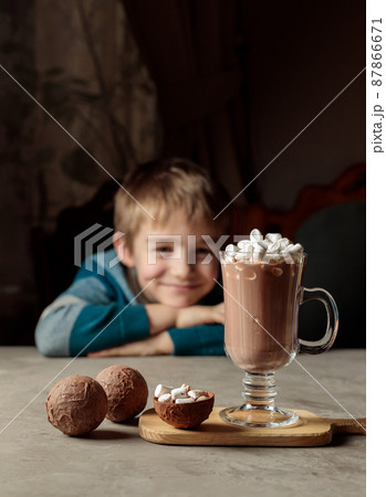 A blond boy with a smile looks at a glass of cocoa or hot chocolate with marshmallows. Chocolate bombs for making cocoa. Selective focus 87866671