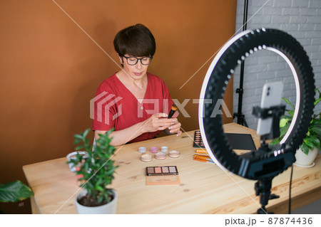 Middle-aged woman talking on cosmetics with makeup eye shadows and blush palette and brushes while recording her video. Mature female making video for her blog on cosmetics Middle-aged woman talking on cosmetics with makeup eye shadows and blush palette and brushes while recording her video. Mature female making video for her blog on cosmetics 87874436