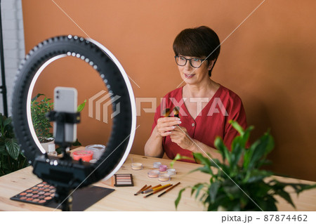 Middle-aged female beauty blogger or influencer with makeup product recording video in room at home. Using ring lamp and smartphone 87874462