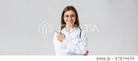 Healthcare workers, medicine and covid-19 pandemic concept. Good-looking professional female doctor in white scrubs and glasses, pointing fingers left, smiling friendly, showing information Healthcare workers, medicine and covid-19 pandemic concept. Good-looking professional female doctor in white scrubs and glasses, pointing fingers left, smiling friendly, showing information 87874634