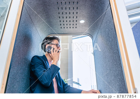 フリーアドレスオフィスの防音電話ボックスで電話をする男性 87877854