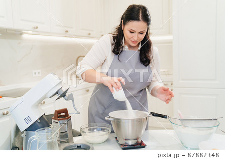 A woman cook adds a sugar to prepare dough or cream in a mixer bowl. A woman cook adds a sugar to prepare dough or cream in a mixer bowl. 87882348