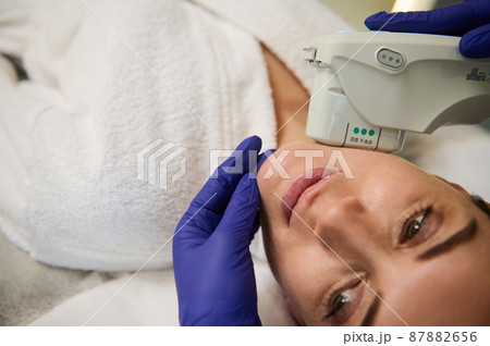 Close-up of the face of a young beautiful woman receiving a facial treatment in the spa. Ultrasonic cleaning and resurfacing of the face with a modern laser device in cosmetology Close-up of the face of a young beautiful woman receiving a facial treatment in the spa. Ultrasonic cleaning and resurfacing of the face with a modern laser device in cosmetology 87882656