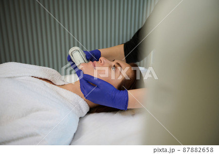Side view of a beautiful woman lying on a massage table during beauty procedures on her face with a modern ultrasonic or laser equipment in a contemporary wellness spa clinic. Health care and medicine Side view of a beautiful woman lying on a massage table during beauty procedures on her face with a modern ultrasonic or laser equipment in a contemporary wellness spa clinic. Health care and medicine 87882658