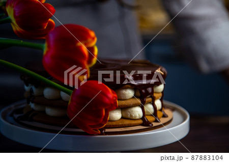 A cake with honey cakes and white cream stands on a white stand and is poured with chocolate on top, next to it are tulips, all in warm colors. High quality photo 87883104
