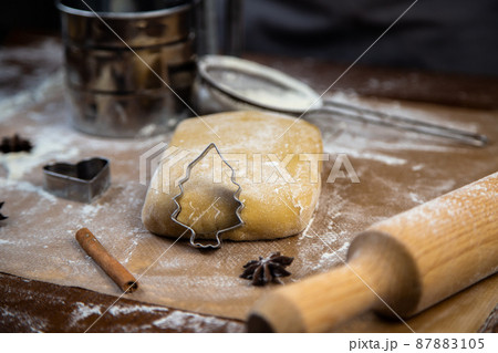 A cookie cutter in the form of a Christmas tree is leaning on the dough, cinnamon and cloves are lying next to it, flour, other cookie cutters and a rolling pin for dough are visible in the background 87883105