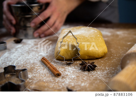 A cookie cutter in the form of a Christmas tree is leaning against the dough, a photo in a dark key, other molds are visible, a rolling pin, an iron sifter for flour, which is held in the hands of a 87883106