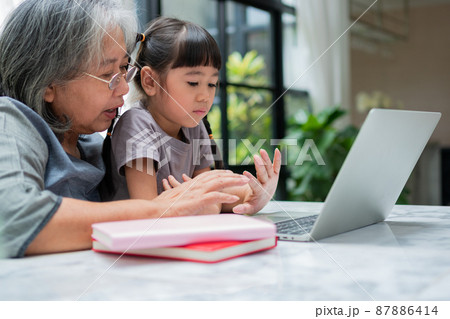 Asian Grandmother with her two grandchildren having fun and playing education games online with a computer notebook at home in the living room. Concept of online education and caring from parents. Asian Grandmother with her two grandchildren having fun and playing education games online with a computer notebook at home in the living room. Concept of online education and caring from parents. 87886414
