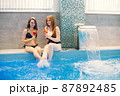Two girls in a black swimwear sitting near swimming pool inside 87892485