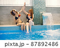 Two girls in a black swimwear sitting near swimming pool inside 87892486
