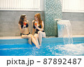 Two girls in a black swimwear sitting near swimming pool inside 87892487