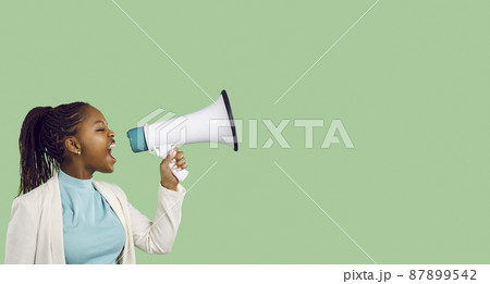 Confident beautiful young black woman holding and yelling through megaphone. Loud activist girl makes important news announcement standing on blank green colour text copyspace studio banner background Confident beautiful young black woman holding and yelling through megaphone. Loud activist girl makes important news announcement standing on blank green colour text copyspace studio banner background 87899542