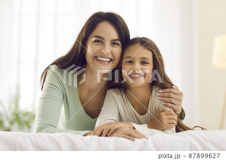 Portrait of happy mother and child in bedroom. Beautiful young mum together with her pretty little daughter cuddling on cosy, warm bed and smiling at camera. Concept of family, love and care Portrait of happy mother and child in bedroom. Beautiful young mum together with her pretty little daughter cuddling on cosy, warm bed and smiling at camera. Concept of family, love and care 87899627