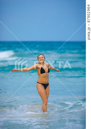 A young girl in a green bathing suit stands by the sea and laughs with her arms outstretched 87902964