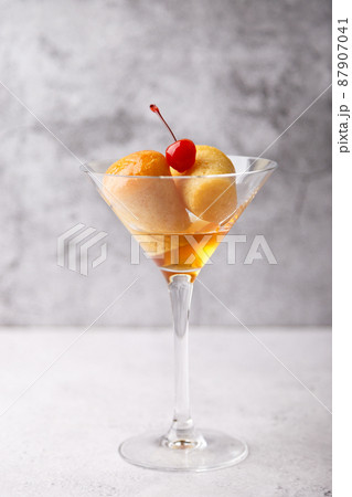 Neapolitan Rum baba (or baba au rum) in a martini glass with a cocktail cherry on a gray background. Small yeast cakes soaked in rum syrup. Traditional Italian pastry. Close-up, selective focus. Neapolitan Rum baba (or baba au rum) in a martini glass with a cocktail cherry on a gray background. Small yeast cakes soaked in rum syrup. Traditional Italian pastry. Close-up, selective focus. 87907041