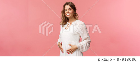 Maternity, women and beauty concept. Tender, cute smiling blond woman in white dress feeling happiness and love, touching belly as expecting child, being pregnant, pink background 87913086
