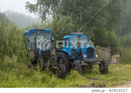 Old blue agricultural tractors on the side of the road near bushes and trees 87914527