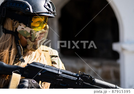 Caucasian woman in a protective suit with a machine gun. A female soldier in a camouflage uniform holds a weapon. 87915395