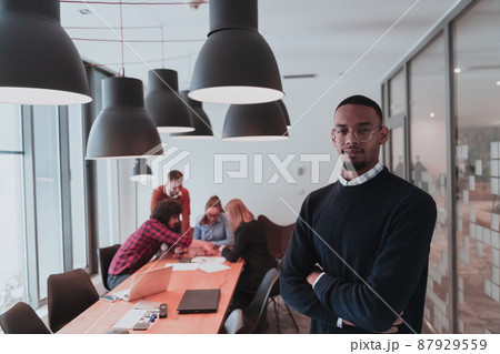 Portrait of happy Afro American millennial male business owner in modern office. Businessman wearing glasses, smiling and looking at camera. Busy diverse team working in background. Leadership concept 87929559