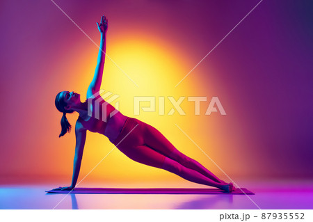 Portrait of young fit girl doing yoga stretching exercises, standing in side plank isolated over gradient pink and yellow background in neon 87935552
