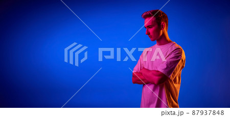 Portrait of young man in white T-shirt posing isolated over blue studio background in pink neon light. Flyer Portrait of young man in white T-shirt posing isolated over blue studio background in pink neon light. Flyer 87937848