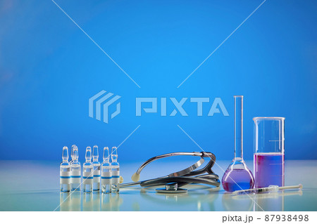 Beaker, medical flask with purple chemical reagent. Six ampoules with liquid, syringe and phonendoscope, blue background. Worldwide pandemic COVID-19 Beaker, medical flask with purple chemical reagent. Six ampoules with liquid, syringe and phonendoscope, blue background. Worldwide pandemic COVID-19 87938498