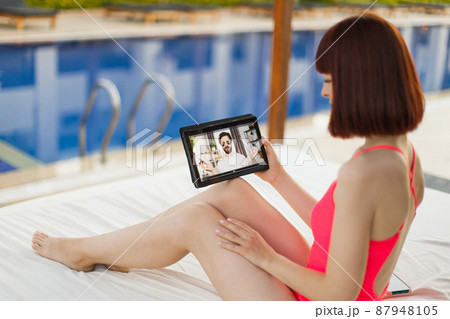Woman in swimsuit, relaxing at resort pool with digital tablet, having a video call with her friend Woman in swimsuit, relaxing at resort pool with digital tablet, having a video call with her friend 87948105