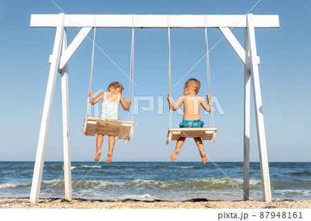 Back view couple of cute little adorable small caucasian sibling kids enjoy having fun swinging and playing at playground sea sand beach against blue ocean water wave and clear sky. Family vacation 87948161