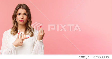 Close-up portrait sad and frustrated young blond girl waited for proposal during romantic date, pointing at finger without weddint ring with puzzled upset expression, standing pink background 87954139