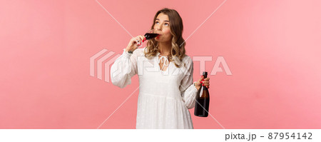 Holidays, spring and party concept. Portrait of young elegant happy blond woman, wear white trendy dress, drinking wine from glass looking up pleased, tasting good drink, hold bottle Holidays, spring and party concept. Portrait of young elegant happy blond woman, wear white trendy dress, drinking wine from glass looking up pleased, tasting good drink, hold bottle 87954142