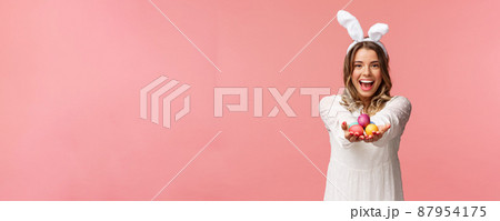 Holidays, spring and party concept. Portrait of lovely, romantic young blond woman in rabbit ears and white dress, giving you painted easter eggs as celebrating orthodox holiday, pink background 87954175