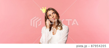 Spring, happiness and celebration concept. Close-up portrait of thoughtful young confident and attractive girl feeling like queen of night party, daydreaming, thinking with crown mask on head 87954205