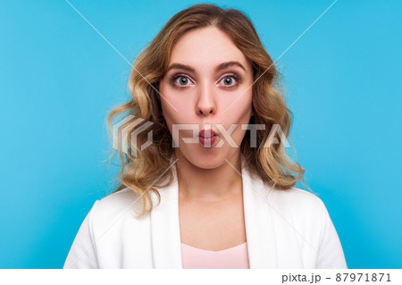 Portrait of funny beautiful woman with wavy hair in white jacket making fish face with pout lips and amazed big eyes, surprised goofy expression. indoor studio shot isolated on blue background 87971871