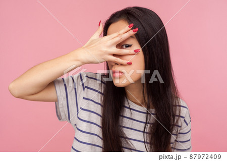 Portrait of nosy curious girl with brunette hair in striped t-shirt peeking through fingers, watching secrets, rumors, spying with inquisitive glance. indoor studio shot isolated on pink background 87972409