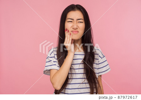 Oral health. Portrait of girl with brunette hair in striped t-shirt touching chin, feeling toothache and grimacing in horrible suffer, dental problems. indoor studio shot isolated on pink background 87972617