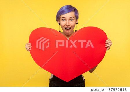 Romantic feelings. Portrait of surprised hipster girl with violet short hair in overalls holding big paper heart and looking with amazed eyes, valentines day. isolated on yellow background studio shot Romantic feelings. Portrait of surprised hipster girl with violet short hair in overalls holding big paper heart and looking with amazed eyes, valentines day. isolated on yellow background studio shot 87972618