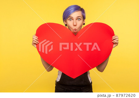 Portrait of funny amusing hipster girl with violet short hair in overalls holding big paper heart and sticking out rolled tongue, comical childish mood, making faces. yellow background studio shot Portrait of funny amusing hipster girl with violet short hair in overalls holding big paper heart and sticking out rolled tongue, comical childish mood, making faces. yellow background studio shot 87972620
