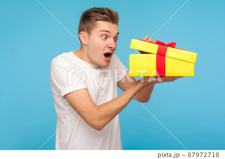 Shocked man in white t-shirt looking inside gift box with curious and surprised expression, opening present before celebration, peeking with amazement. indoor studio shot isolated on blue background 87972718