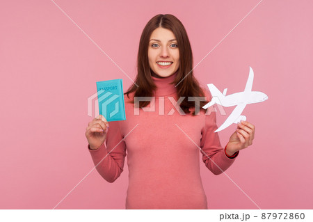 Happy brunette woman in pink sweater holding paper plane model and passport in hands looking at camera with toothy smile. Indoor studio shot isolated on pink background 87972860