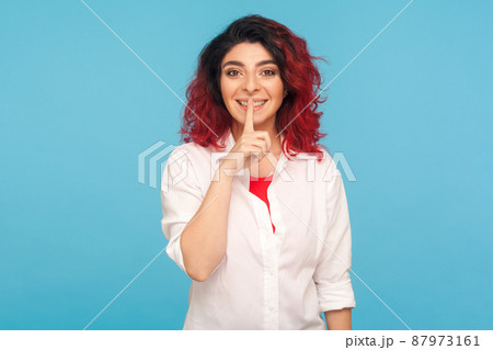 Shh, keep quiet. Portrait of positive hipster woman with fancy red hair in white shirt holding finger on lips, hushing and showing silence gesture. indoor studio shot isolated on blue background 87973161