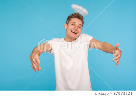 Let me embrace you. Angelic kind-hearted man with halo above head outstretching hands to hug and looking at camera with childish joyful expression. indoor studio shot isolated on blue background Let me embrace you. Angelic kind-hearted man with halo above head outstretching hands to hug and looking at camera with childish joyful expression. indoor studio shot isolated on blue background 87973221