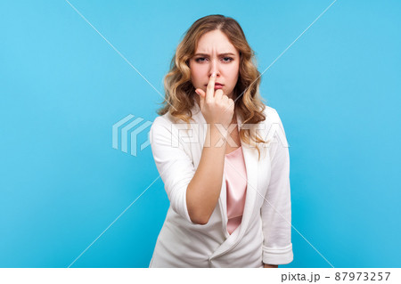 You are liar. Portrait of upset woman with wavy hair in white jacket touching nose showing lie gesture and frowning face, having suspicious about falsehood, dishonesty. studio shot, blue background You are liar. Portrait of upset woman with wavy hair in white jacket touching nose showing lie gesture and frowning face, having suspicious about falsehood, dishonesty. studio shot, blue background 87973257