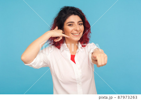Hey you, call me back. Happy woman with fancy red hair making telephone gesture near head and winking playfully, waiting for call, wants to talk on phone. studio shot isolated on blue background 87973263