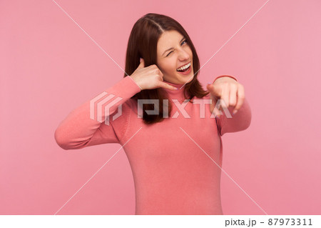 Communicative positive brunette woman showing call gesture and pointing finger at you, waiting for your call. Indoor studio shot isolated on pink background 87973311
