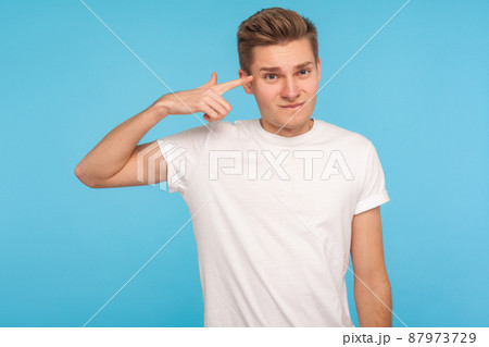 You are idiot. Displeased man in casual white t-shirt holding finger against his temple, making crazy stupid gesture, blaming in senseless dumb talk. indoor studio shot isolated on blue background 87973729