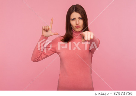 You are loser. Bossy strict woman with brown hair pointing finger at you showing loser gesture, blaming and mocking, social pressure. Indoor studio shot isolated on pink background 87973756